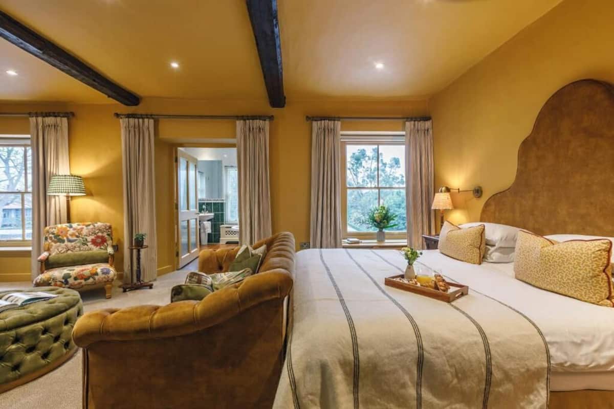 Quarto de hotel com uma cama grande, um sofá, uma cadeira e uma mesa pequena. Há cortinas nas janelas e um abajur na mesinha de cabeceira.