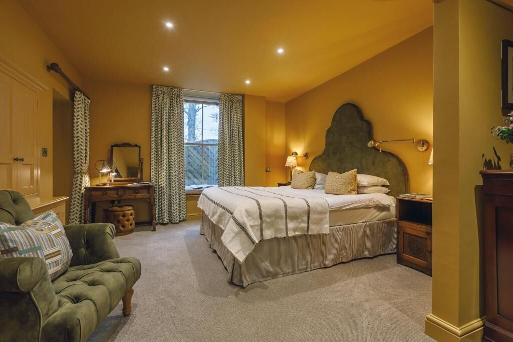 Un dormitorio con una cama, un sillón verde, una mesita de noche, una lámpara, un ventanal con cortinas y un espejo.