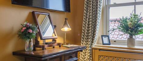 A room with a wooden desk, a mirror, a lamp, a vase with flowers, a framed picture, and a window with curtains.