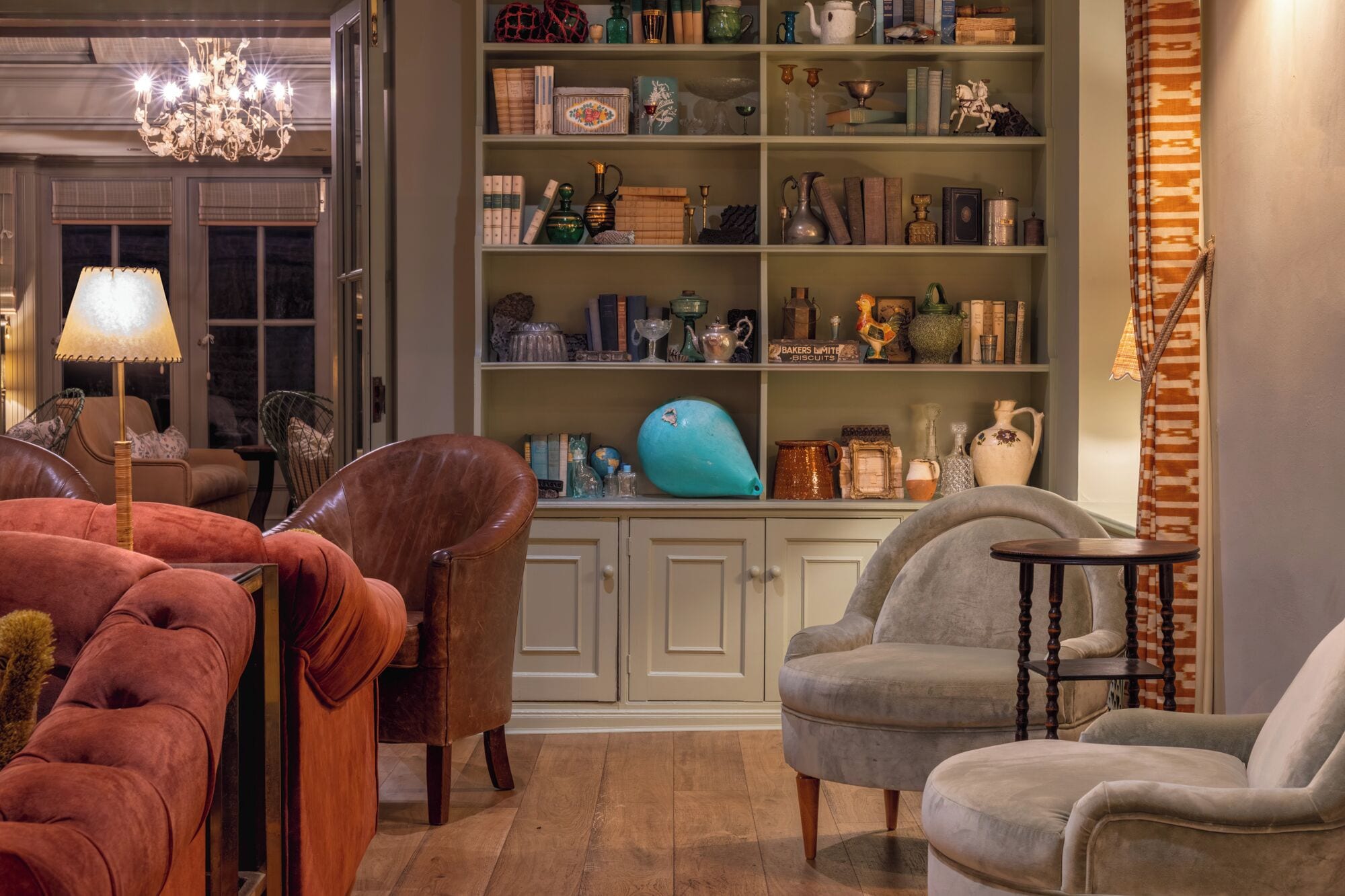 A cozy living room with a bookshelf filled with various decorative items and furniture including a brown leather sofa, a grey armchair, and a small round table.
