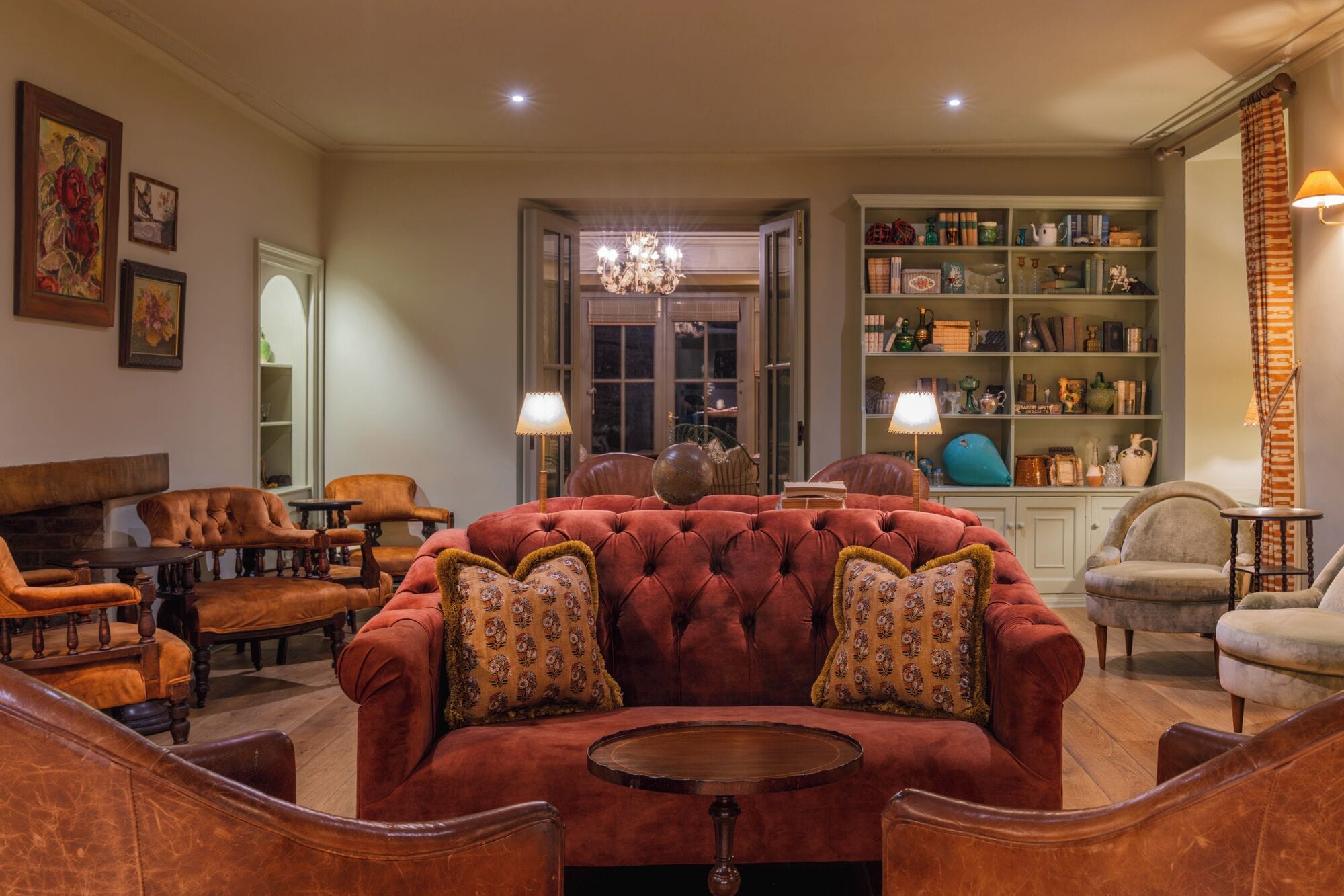 Un vaste salon agrémenté d’un grand canapé rouge, de plusieurs fauteuils et d’une table basse en bois. On y trouve une bibliothèque remplie de livres et d’objets décoratifs, ainsi qu’un lustre suspendu au plafond.