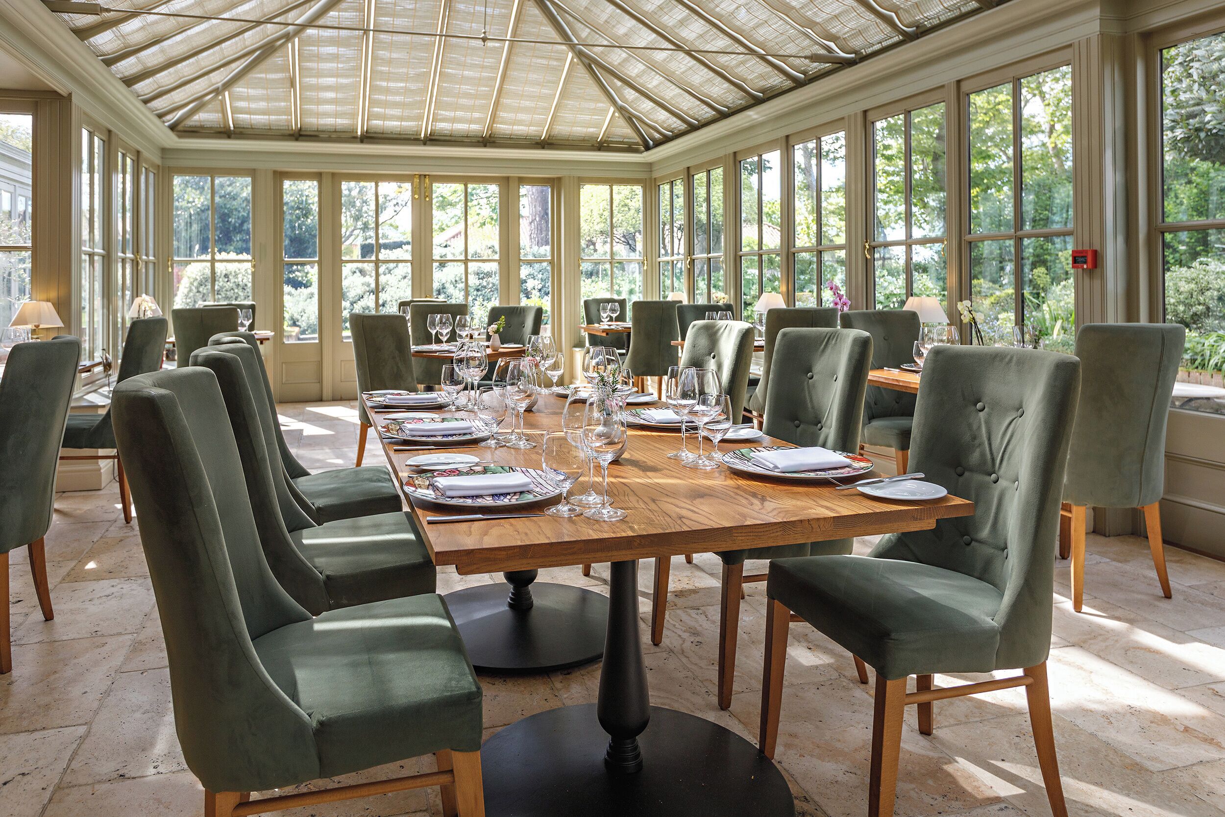A conservatory dining area with a long table set for a meal, surrounded by green upholstered chairs.