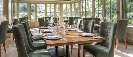 A conservatory dining area with a long table set for a meal, surrounded by green upholstered chairs.