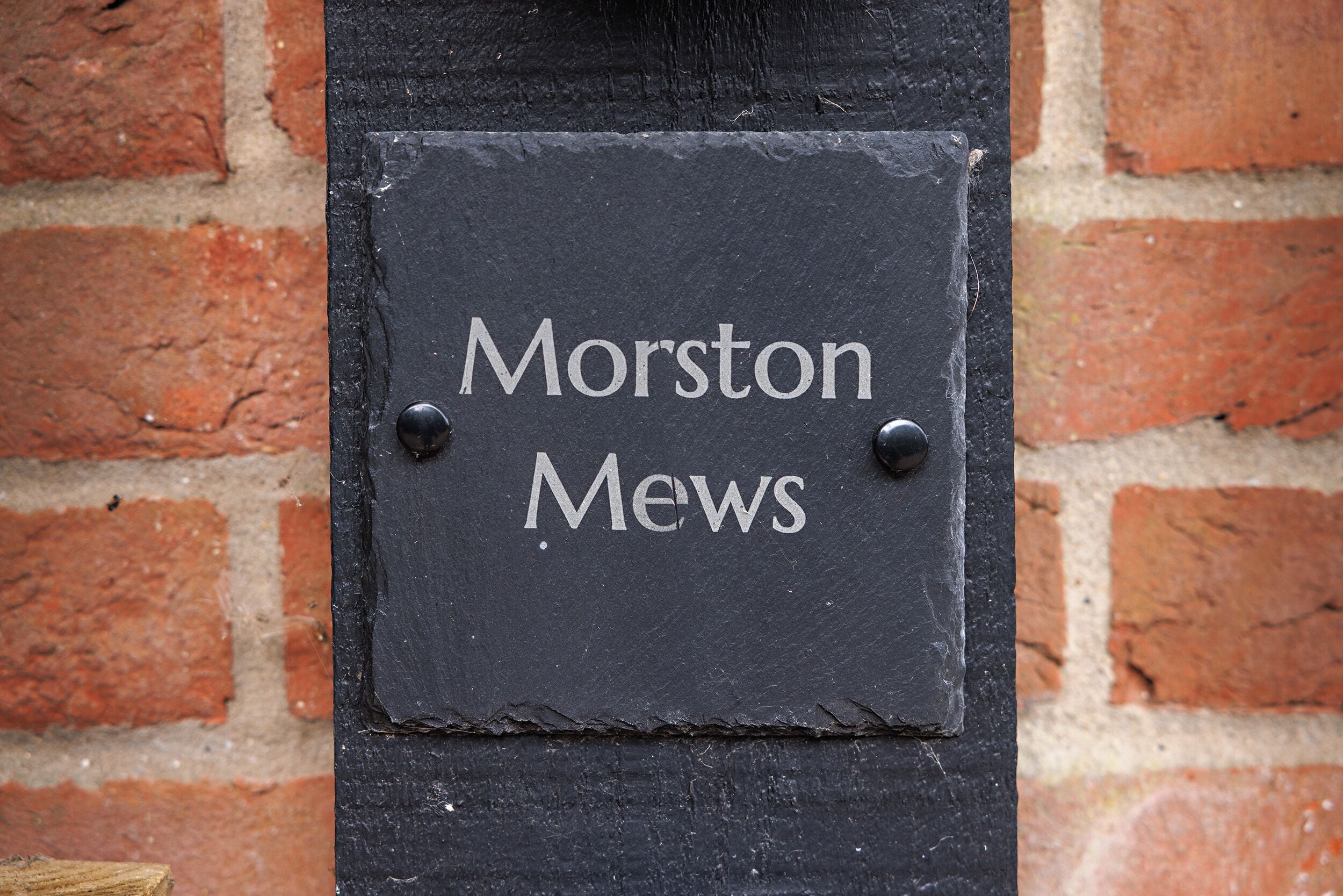 Un letrero de pizarra negra con letras blancas que dice 'Morston Mews' fijado a una pared de ladrillo.