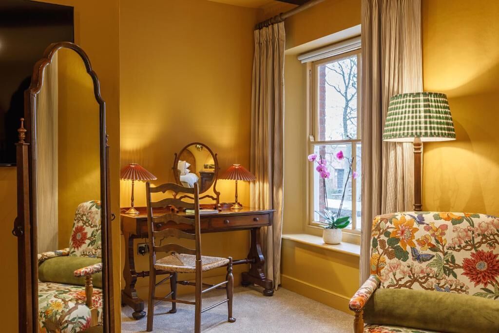 A room with a wooden desk, a chair, a mirror, a lamp, and a floral armchair.