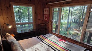 Ein Zimmer in einer Holzhütte mit einem Bett, einem Schreibtisch, einem Sessel und großen Fenstern mit Blick auf den Wald.