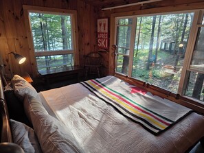 A wooden cabin room with a bed, a desk, a chair, and large windows offering a forest view. - Red pines lake house,  great for 2 families,Pet-friendly.  Beautiful sunsets! (Highlands East)