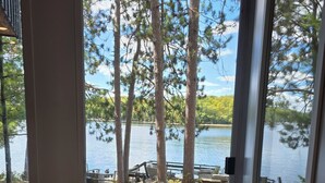 Blick durch eine Glastür auf einen Deckbereich mit Grill, Holzmöbeln und einen See mit Bäumen.