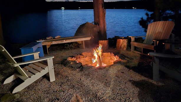 Ein Lagerfeuer am Seeufer mit zwei Adirondack-Stühlen, einer Holzbank und einem Baum.