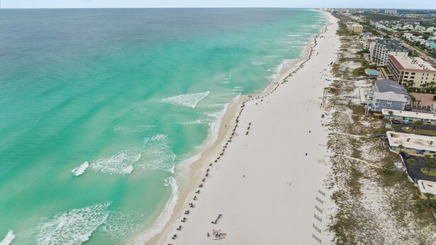 Vista aérea de una playa con una extensa franja de arena, algunos edificios y cielo despejado.