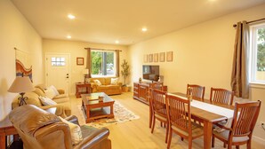 A spacious living room with a sofa, coffee table, dining table, and chairs. There is a television and a window with curtains. - Townhouse on the Trail (Sequim)