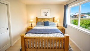 A bedroom with a wooden bed, a headboard with a picture, bedside lamps, and a window with a view of houses. - Townhouse on the Trail (Sequim)