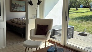 Een moderne woonkamer met een bank, fauteuil en een houten salontafel. Aan de muur hangt een schilderij, er staat een lamp en er is uitzicht op het landschap door grote schuifdeuren.