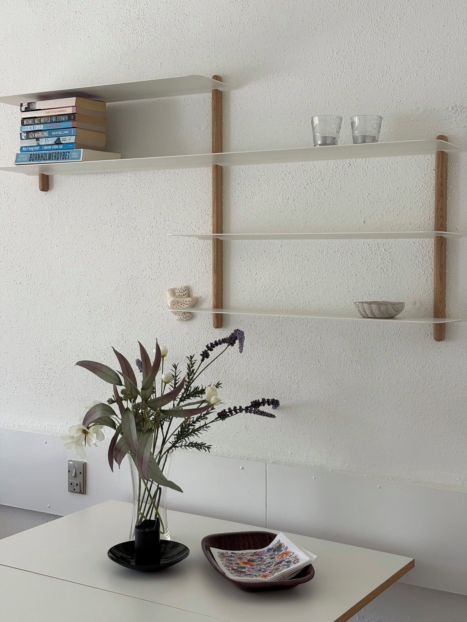 A white table with a vase of flowers, a black bowl, and a stack of magazines. A white shelf with books and glasses is mounted on the wall.