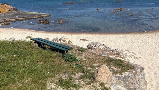 Een strand met een betonnen pier, een groene bank en helderblauw water.