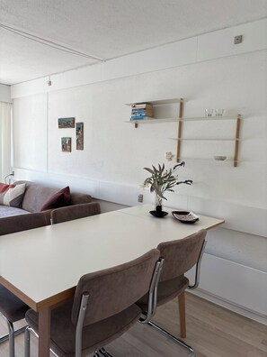 A modern dining area with a white table, brown chairs, a sofa, and wall-mounted shelves with books and decorative items. - Lovely holiday apartment near centre (Allinge)