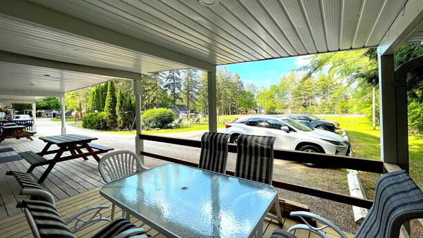 A covered outdoor seating area with a glass table and chairs, overlooking a parking lot and greenery. - Bonnie 1 Cottage/Resort/Orillia, Muskoka (Washago)