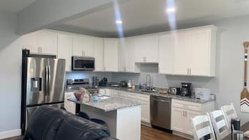 Una cocina moderna con armarios blancos, electrodomésticos de acero inoxidable, encimera de granito y comedor con mesa y sillas de madera.