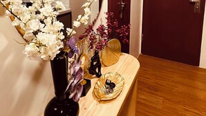 Ein Flur mit einem runden Spiegel, einem Holztischchen mit Blumen in einer Vase, einer kleinen Statue und einer dekorativen Schale. Eine Tür und eine Wandleuchte sind ebenfalls zu sehen.