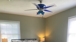 Un dormitorio con cama, mesita de noche, lámpara, ventilador de techo y ventana con persianas.