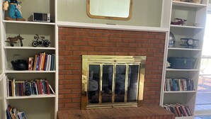Un salón con chimenea de ladrillo, piso de madera y estanterías blancas llenas de libros y objetos decorativos.