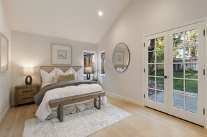 Un dormitorio con cama, mesitas de noche, un banco, un ventanal grande y una puerta que da a un jardín.