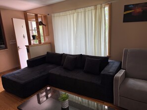 A living room with a sectional sofa, a glass coffee table, a grey armchair, a door with a 'Welcome' sign, and a window with sheer curtains.