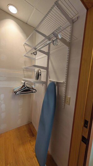 A compact room with a built-in ironing board, a shelf with hanging clothes, and a wall-mounted rack with various items. - 3BR Gilford Condo, Newly Updated, 2 private balconies, 2 min to walk town docks. (Gilford)