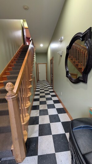 Ein Flur mit einem Schachbrettmuster auf dem Boden, einer Holztreppe, einem Wandspiegel und einer Tür am Ende.