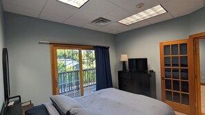 A hotel room with a bed, a TV, a desk, and a balcony with a view of trees and a building.