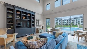 A spacious living room with a large dark blue entertainment unit, a blue sofa with patterned cushions, and a view of a swimming pool and apartment buildings through large windows. - Landing | Beautiful 1BD, Clubhouse (McKinney)