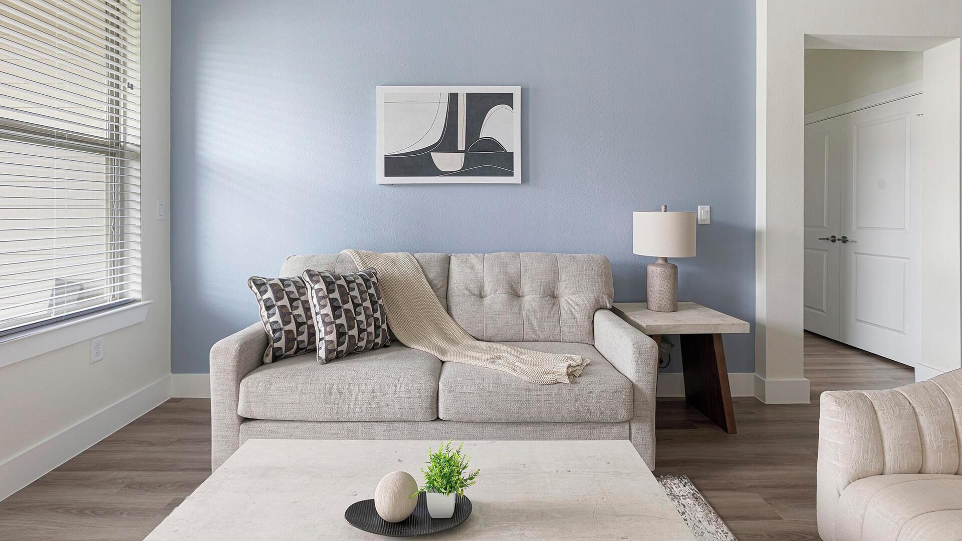A living room with a grey sofa, a side table with a lamp, a framed artwork on the wall, and a coffee table with decorative items.