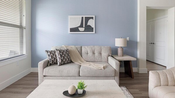 Ein Wohnzimmer mit grauem Sofa, einem Beistelltisch mit Lampe, einem gerahmten Kunstwerk an der Wand und einem Couchtisch mit Dekorationen.