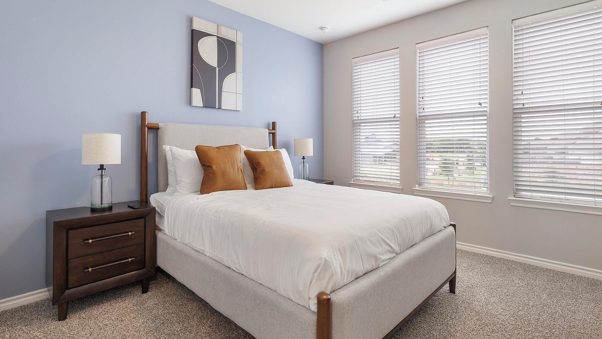 A bedroom with a bed, nightstands, a wall art piece, and large windows with blinds.