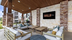 A covered outdoor seating area with sofas, a table, and a TV screen displaying 'OVILLA LEGACY SQUARE'.