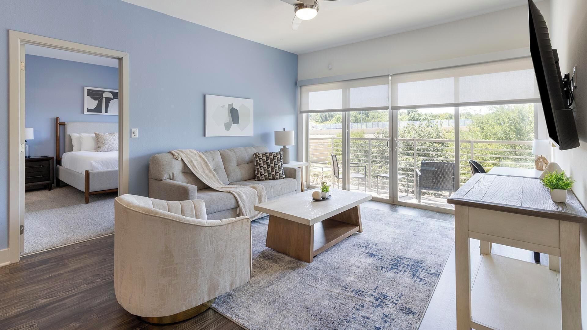 A modern living room with a sofa, coffee table, and a TV. There is a balcony with a view of greenery.
