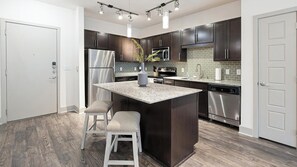 A modern kitchen with dark wood cabinets, stainless steel appliances, a granite countertop island, and bar stools. - Landing | Stylish 1BD, Gym (Dallas)