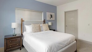 Une chambre à coucher comprenant un lit, des tables de chevet, une fenêtre avec des stores, une porte et une œuvre d’art accrochée au mur.