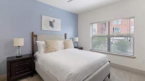 Une chambre à coucher comprenant un lit, deux tables de chevet, une commode, une lampe fixée au mur, une fenêtre avec des stores et un tableau accroché au mur.