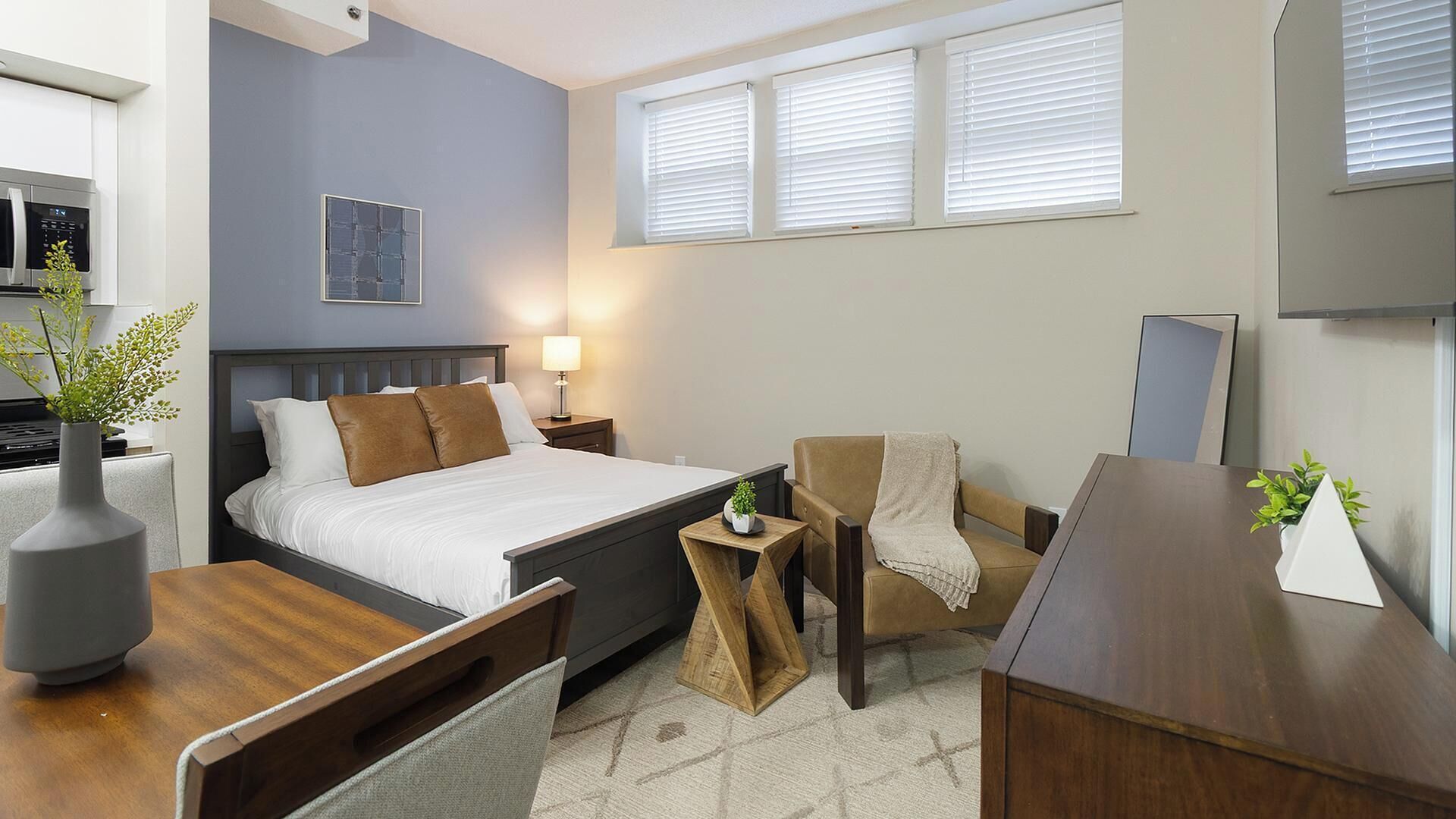 A modern hotel room with a bed, bedside table, chair, desk, and a small table with a plant.