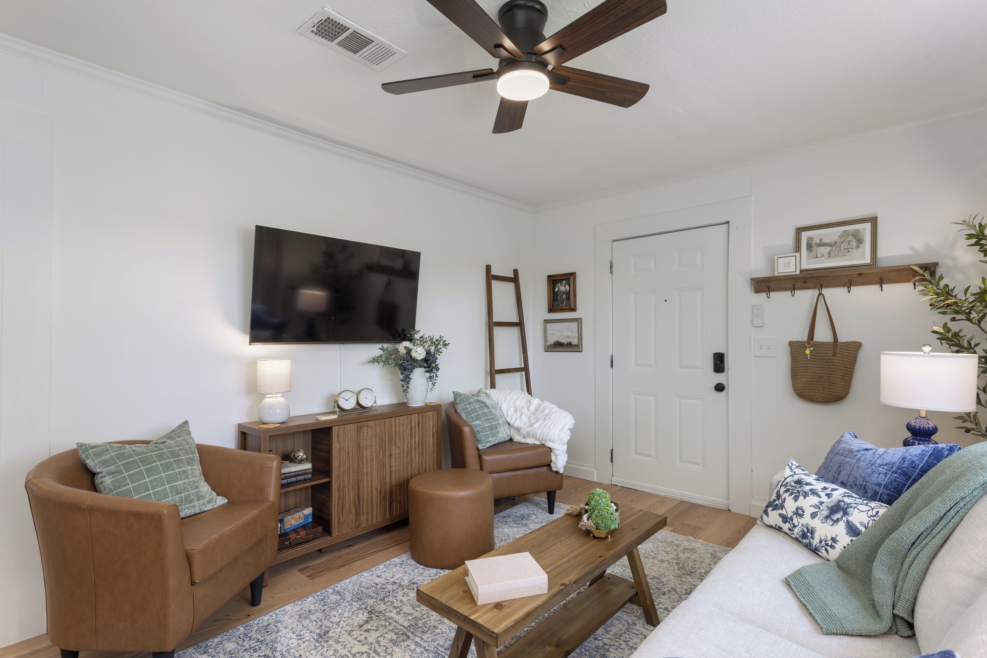 Un salon avec un canapé marron, une table basse en bois, un divan blanc, un ventilateur de plafond et un téléviseur fixé au mur.