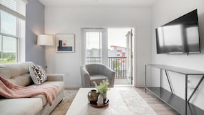 A modern living room with a sofa, armchair, coffee table, TV, and a view through a door to an outdoor balcony. - Landing | Chic 2BD, Gym, Clubhouse (Kissimmee)