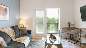 Ein Wohnzimmer mit einer Couch, einem Glastisch, einem Essbereich mit Tisch und Stühlen, einem Balkon mit Geländer und einem an der Wand befestigten Fernseher.