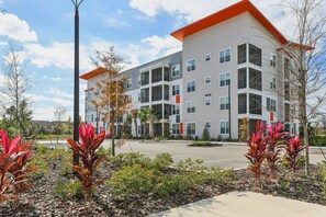 A multi-story building with balconies, a parking lot, and landscaped areas with red-leafed plants. - Landing | Incredible 1BD, Gym, Clubhouse (Kissimmee)
