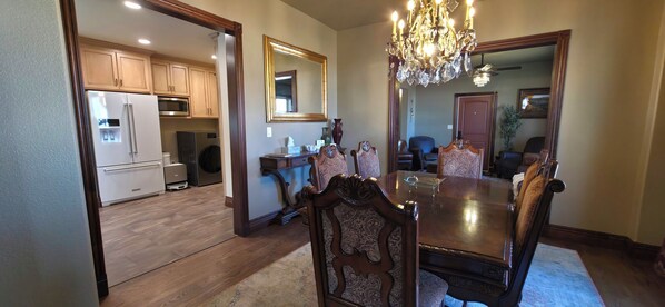 A dining room with a wooden table and chairs, a chandelier, and a kitchen in the background.