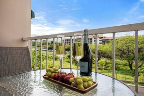 A table with a bottle of wine, two glasses, and a wooden tray with fruits on a balcony with a view of greenery and buildings. - Maui Getaway | Walk to Beach, Pool & Hot Tub (Kihei)