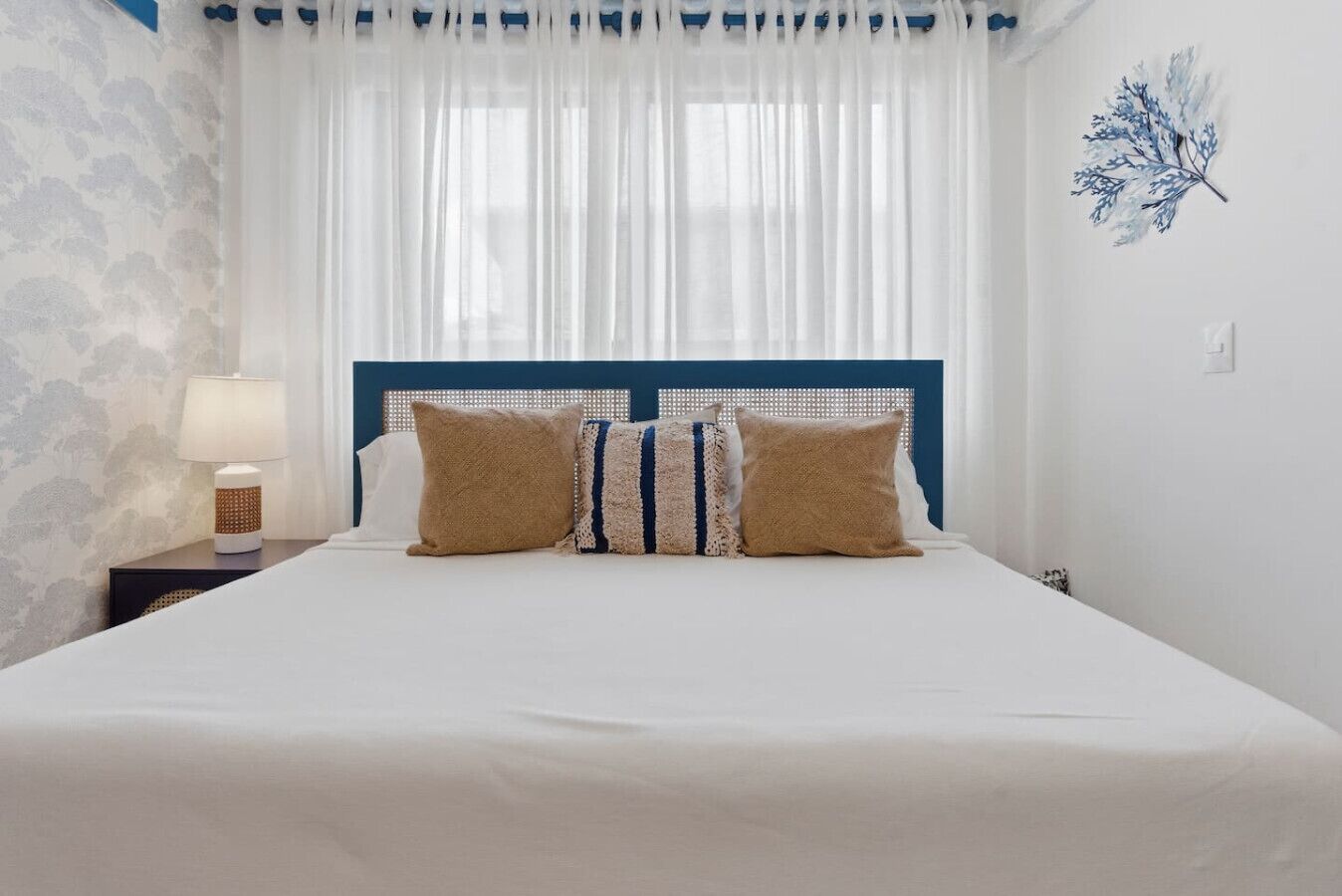 Un dormitorio con cama, cortinas blancas, una lámpara de pared y una hoja decorativa en la pared.