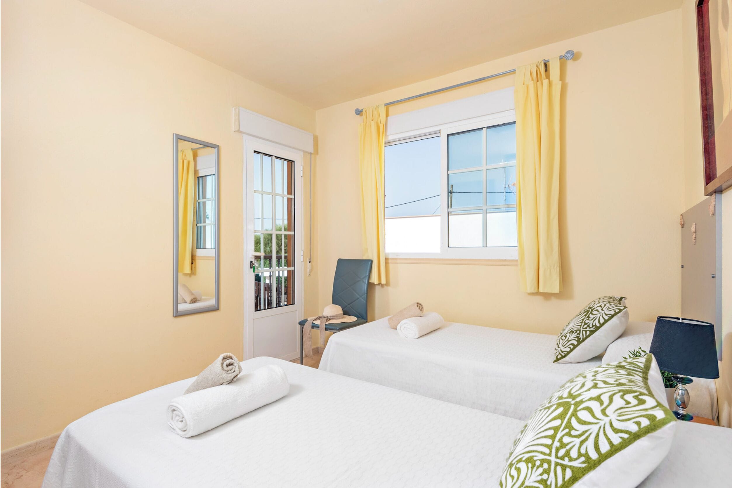 Une chambre d’hôtel avec deux lits, une fenêtre avec des rideaux jaunes, un miroir, une chaise, une table de chevet et une lampe.