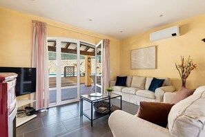 A living room with a sofa, a coffee table, a TV, and a sliding glass door leading to an outdoor area with a pool. - Villa Lamar - Three Bedroom Villa, Sleeps 6 (Alayor)