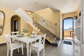 A dining area with a white table set for four, a staircase with a metal railing, and a view of the sea through an open door. - Villa Lamar - Three Bedroom Villa, Sleeps 6 (Alayor)